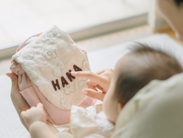 ベビーが「HANA」刺繍を指さして触れる花ピンクのお絵かきステッチリュック-手刺しゅう風の立体感が伝わるアップ